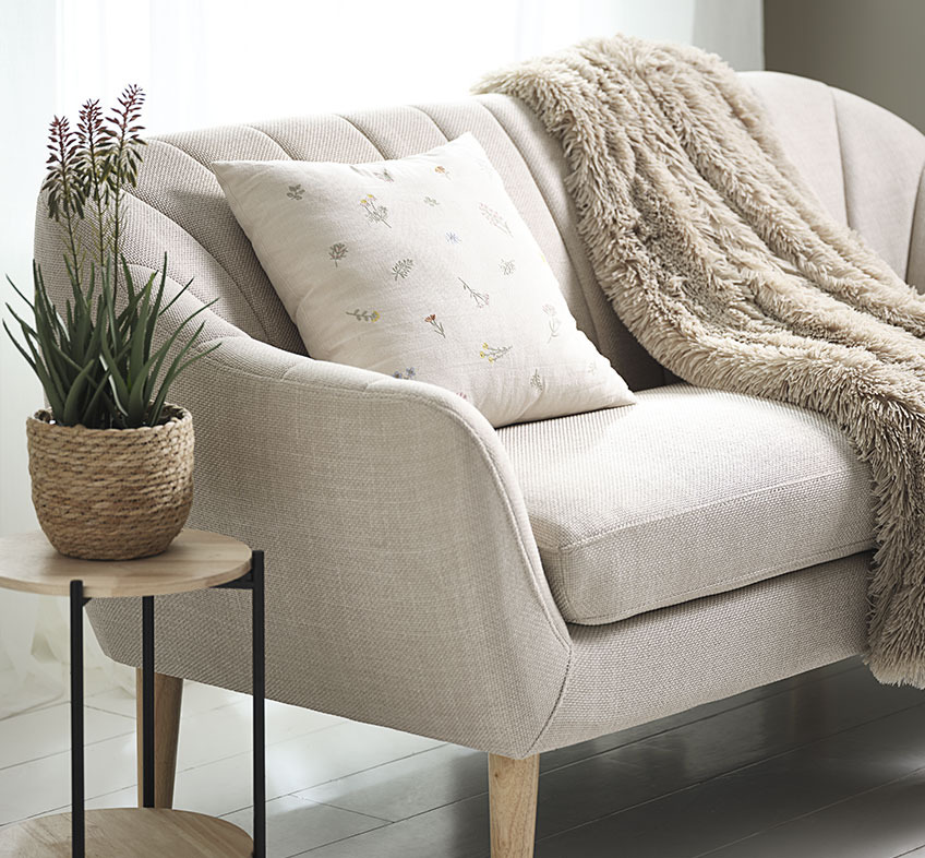 Plante artificielle RASMUS dans le cache-pot STEFAN, coussin AGERTIDSEL et plaid MYRFIOL Plante artificielle d’aloe vera avec fleurs, coussin brodé et plaid en fausse fourrure sur un canapé beige