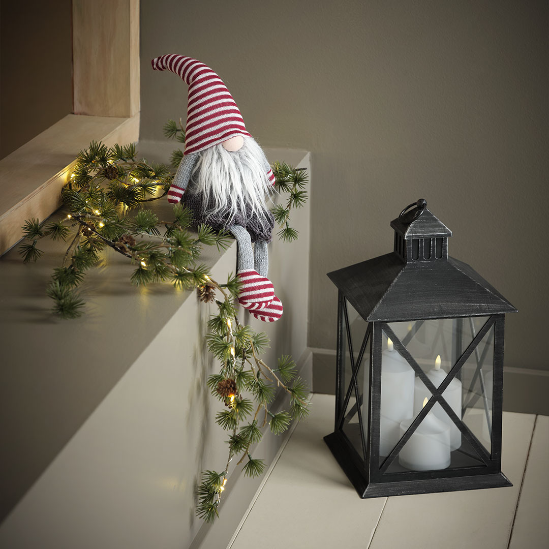 Lutin de Noël SVADILFARI et lanterne PULKA Lutin de Noël avec un bonnet, des gants et des chaussettes à rayures rouges à côté d’une lanterne noire