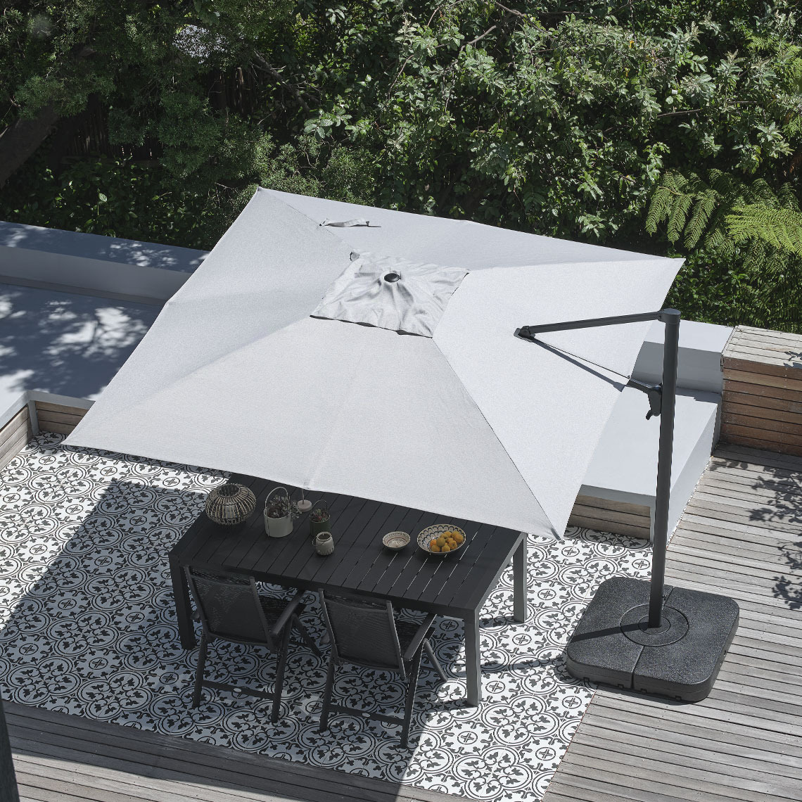 Parasol déporté BUBERG en gris, avec fonction d’inclinaison, manivelle de réglage et fentes de ventilation. Grand parasol déporté gris, de forme carrée, installé au‑dessus d’une table de repas d’extérieur avec des chaises de jardin