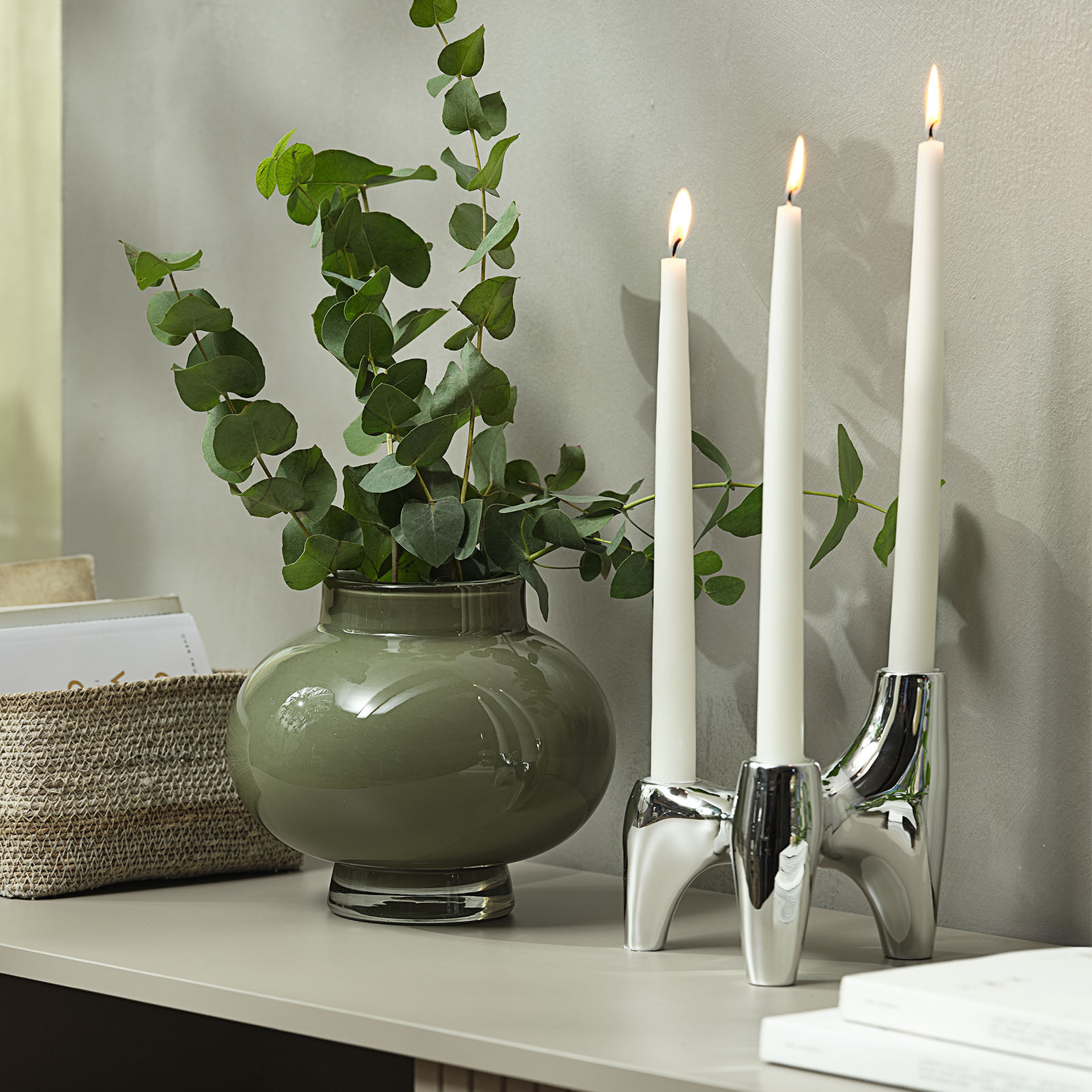Bougeoir effet chrome avec trois bougies à côté d’un vase en verre vert contenant de l’eucalyptus artificiel